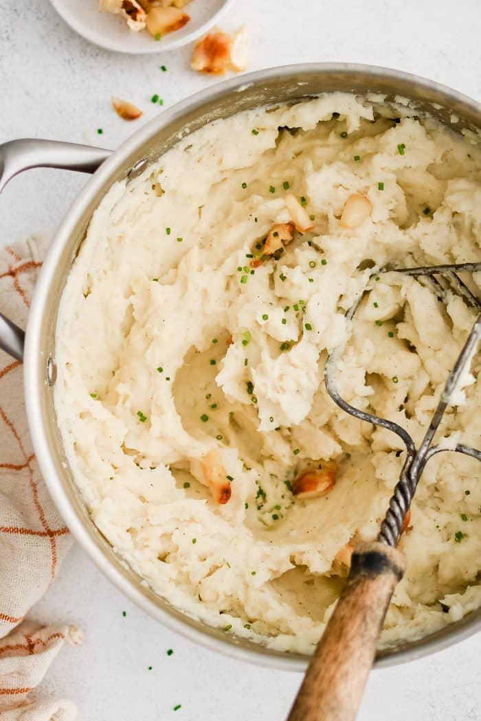 Hand potato masher resting in a large pot filled with creamy mashed russet potatoes with whole cloves of roasted garlic.