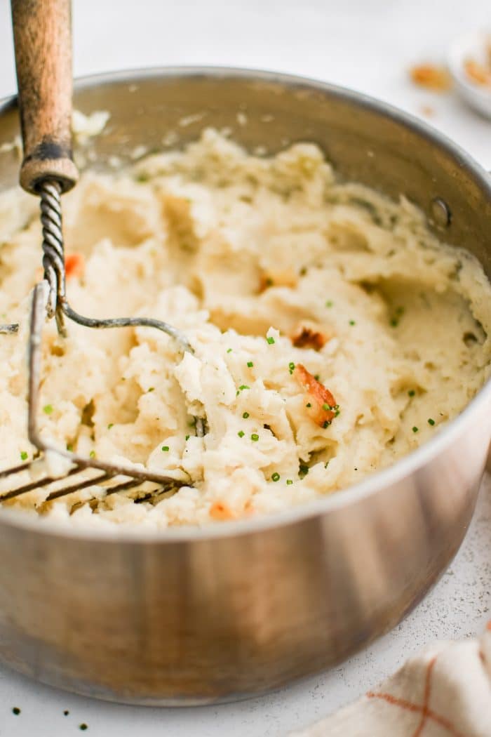 Hand potato masher resting in a large pot filled with creamy mashed russet potatoes with whole cloves of roasted garlic.