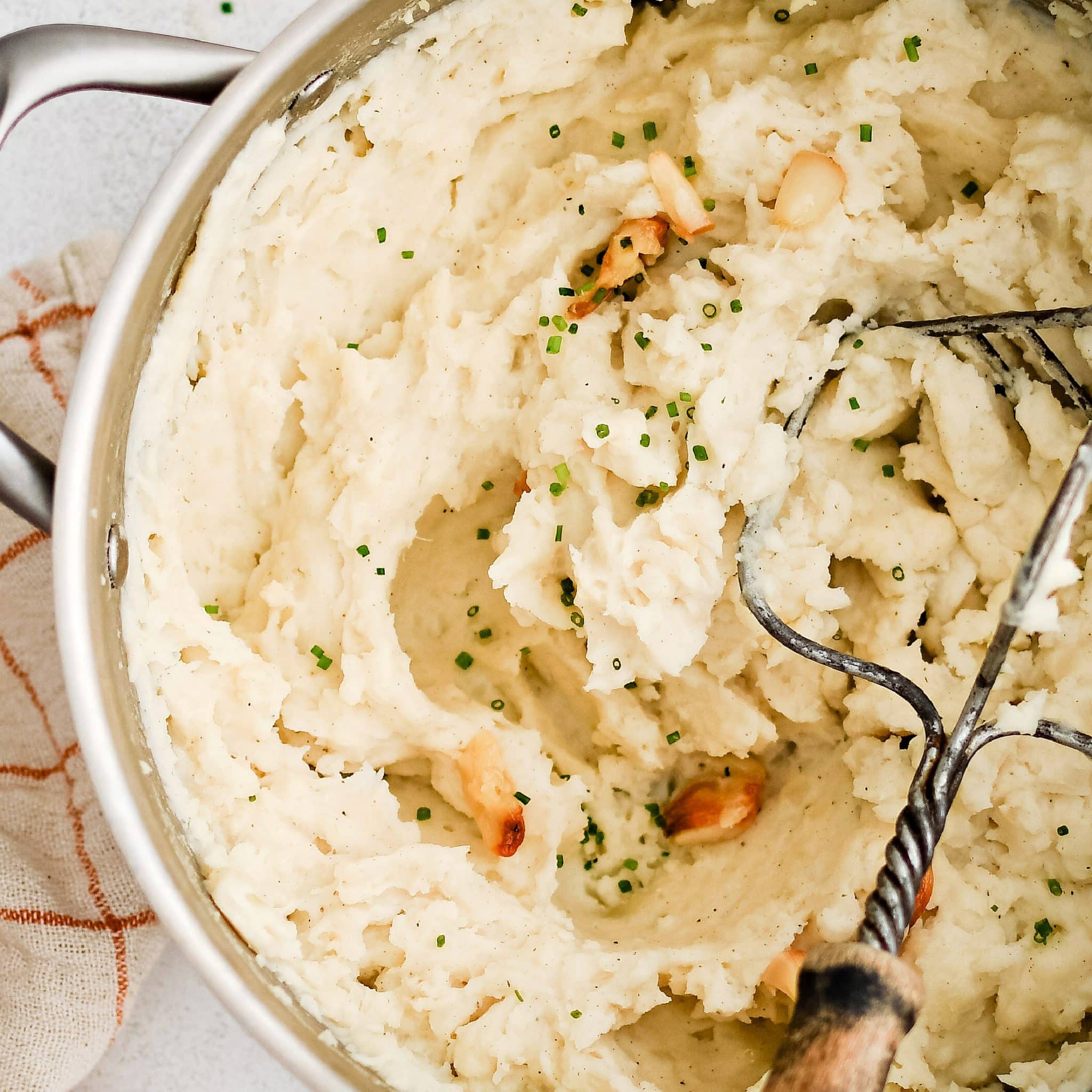 Hand potato masher resting in a large pot filled with creamy mashed russet potatoes with whole cloves of roasted garlic.