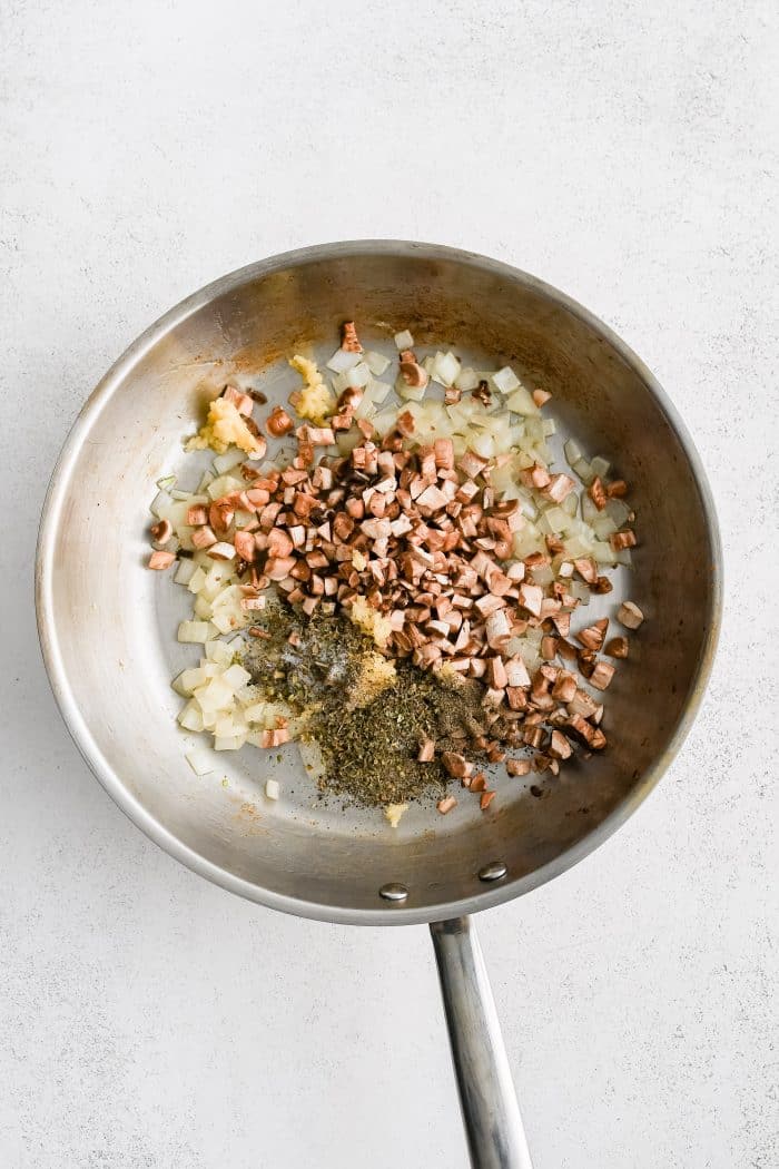 Chopped mushroom stems, Italian seasoning, garlic, salt, and black pepper added to a large stainless steel skillet filled with softened onions.