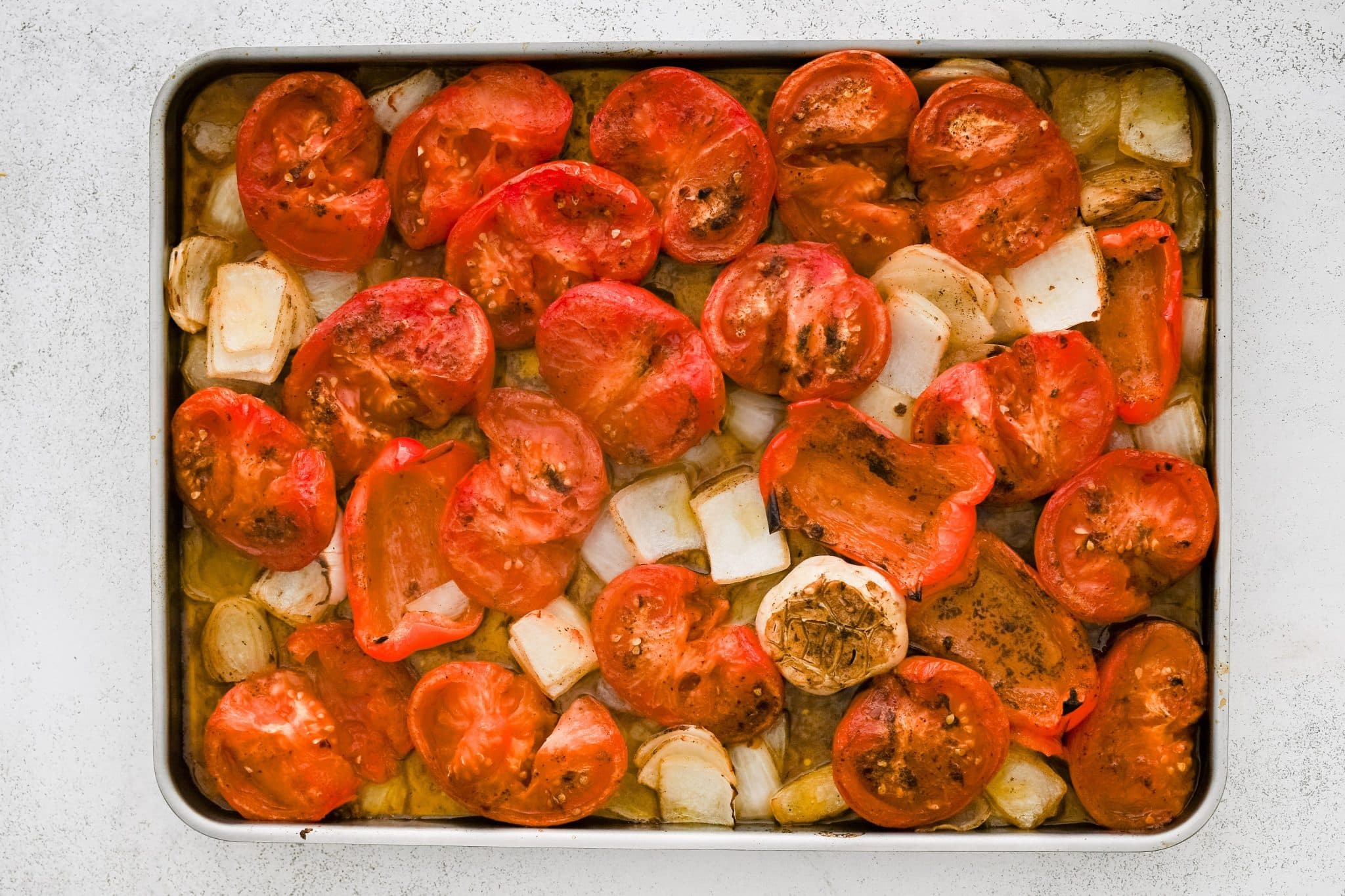 Large rimmed baking sheet filled with roasted halved tomatoes, a bulb of garlic, and roughly chopped onion and red bell pepper.