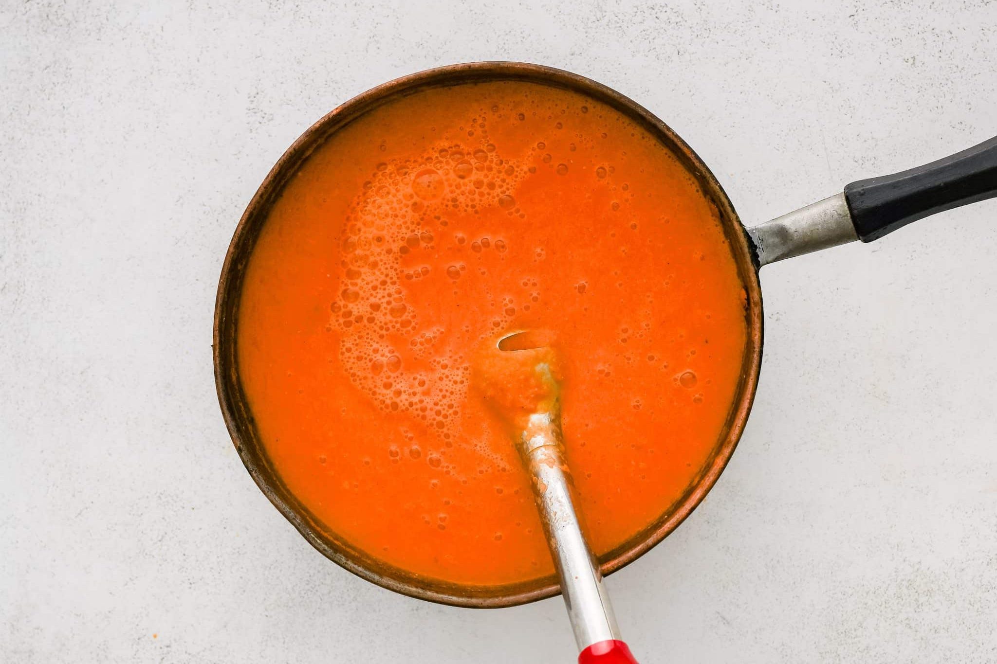 Immersion blender blending roasted tomatoes, garlic, onion, and red bell pepper with broth in a large soup pot.