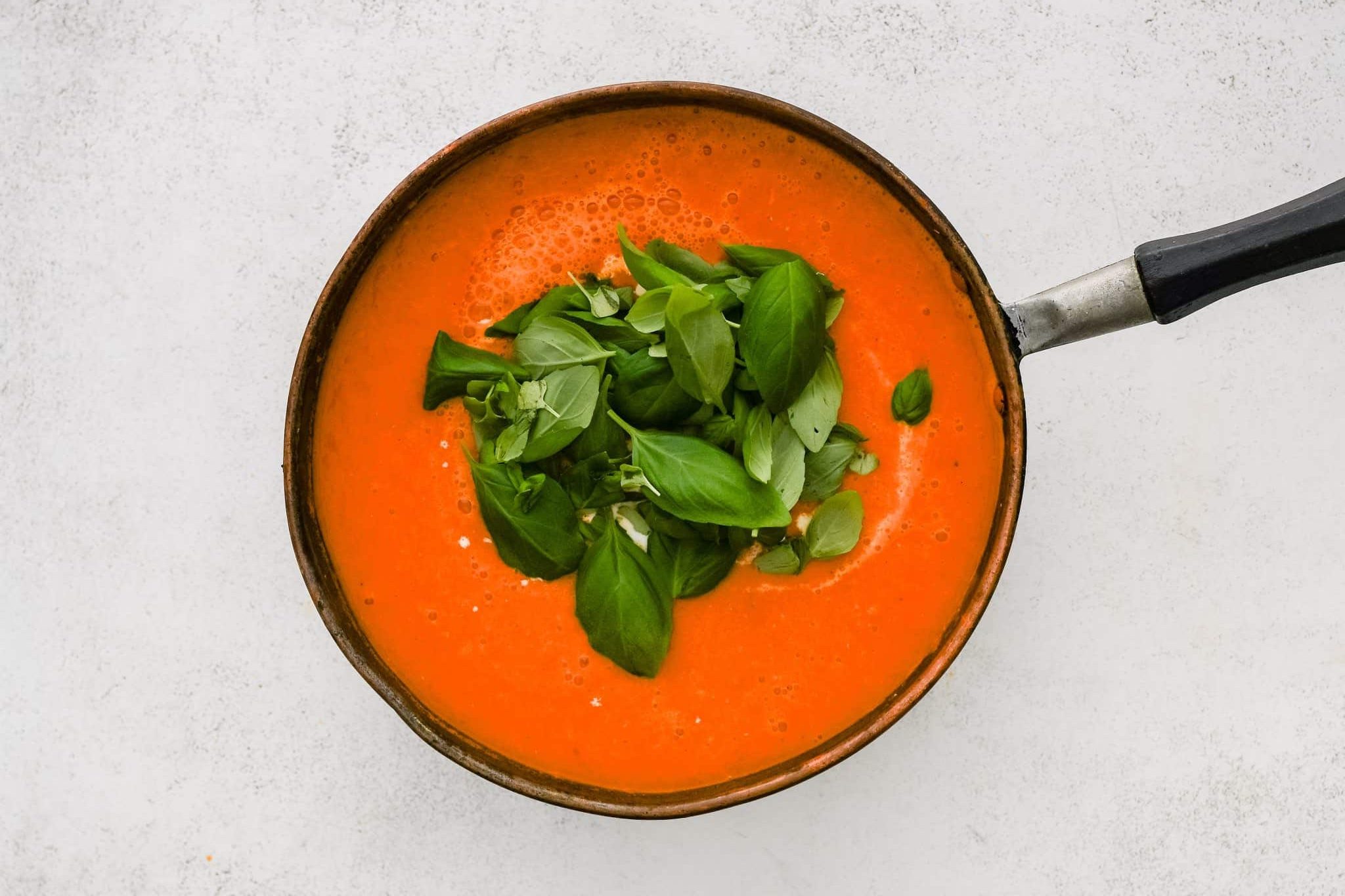 Fresh basil and heavy cream added to a large pot filled with simmering fully blended roasted tomatoes, onion, garlic, and red bell pepper.