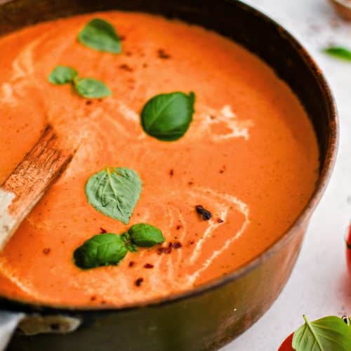 Large soup pot filled with thick, creamy, and rich blended tomato bisque garnished with fresh basil leaves, cracked red pepper, and a drizzle of fresh cream.