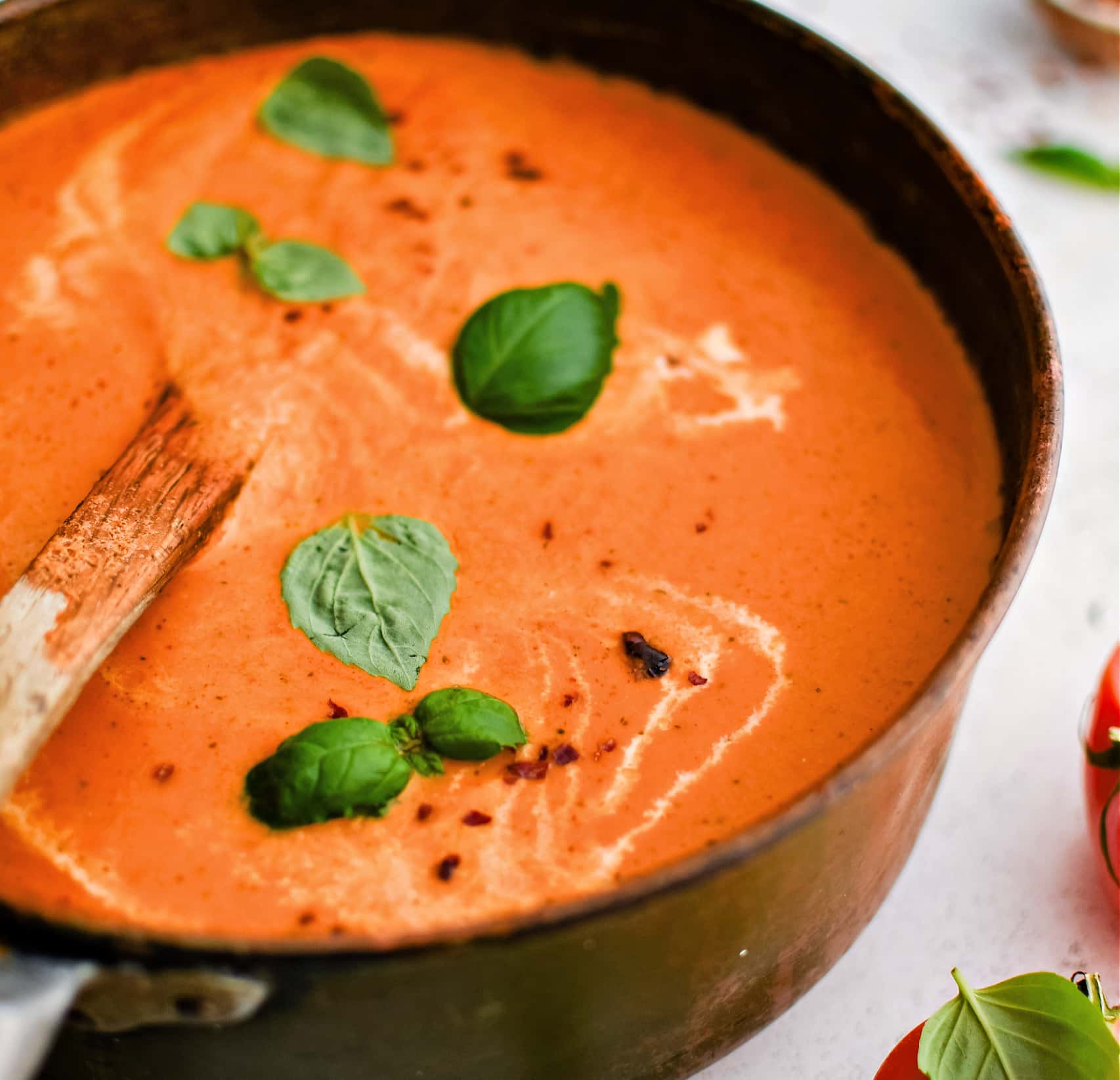 Large soup pot filled with thick, creamy, and rich blended tomato bisque garnished with fresh basil leaves, cracked red pepper, and a drizzle of fresh cream.