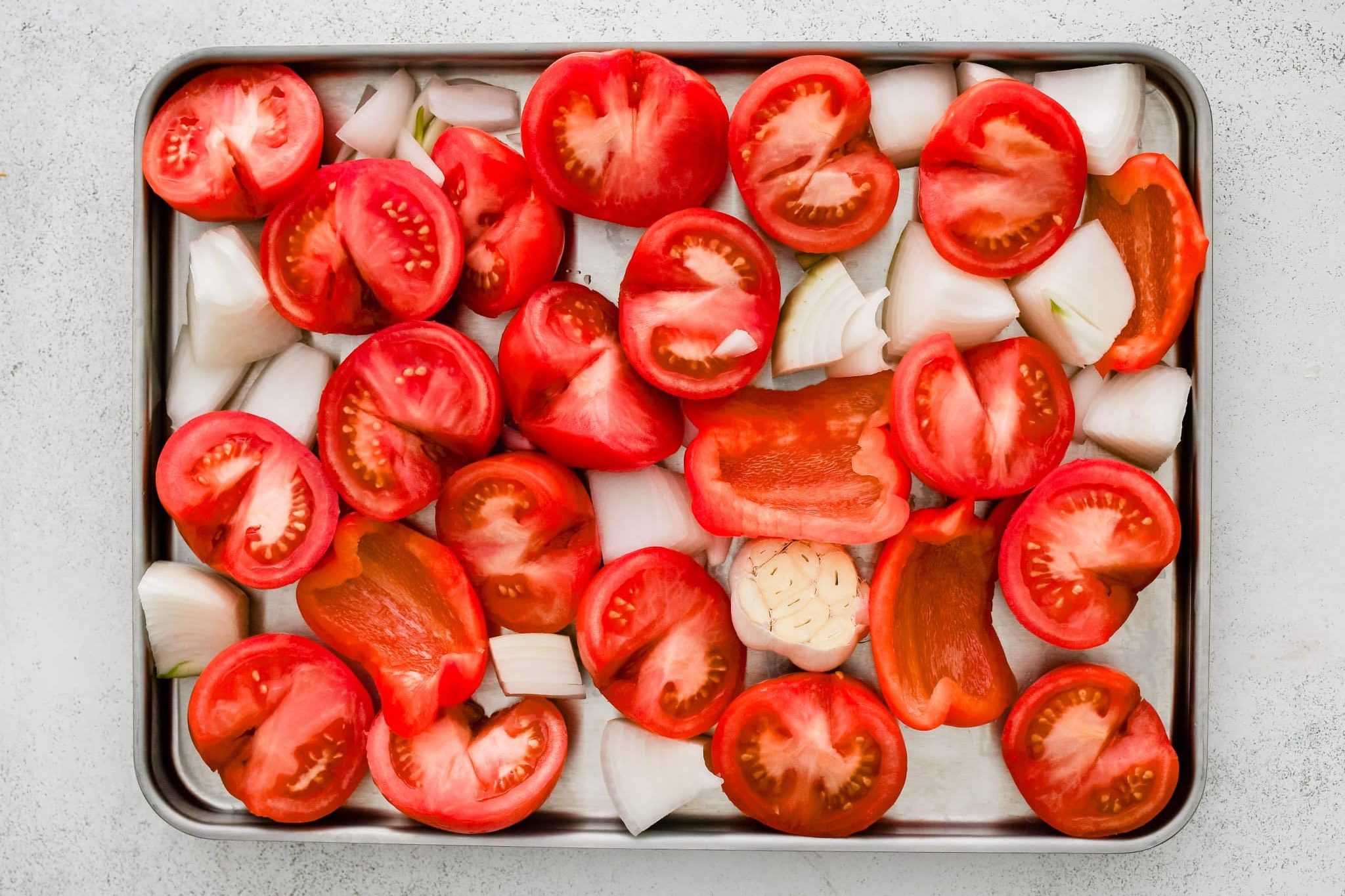 Large rimmed baking sheet filled with halved tomatoes, a bulb of garlic, and roughly chopped onion and red bell pepper.