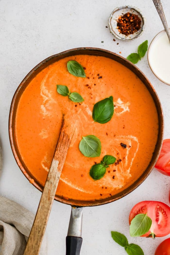 Large soup pot filled with thick, creamy, and rich blended tomato bisque garnished with fresh basil leaves, cracked red pepper, and a drizzle of fresh cream.