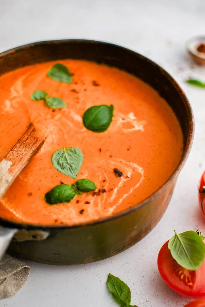 Large soup pot filled with thick, creamy, and rich blended tomato bisque garnished with fresh basil leaves, cracked red pepper, and a drizzle of fresh cream.