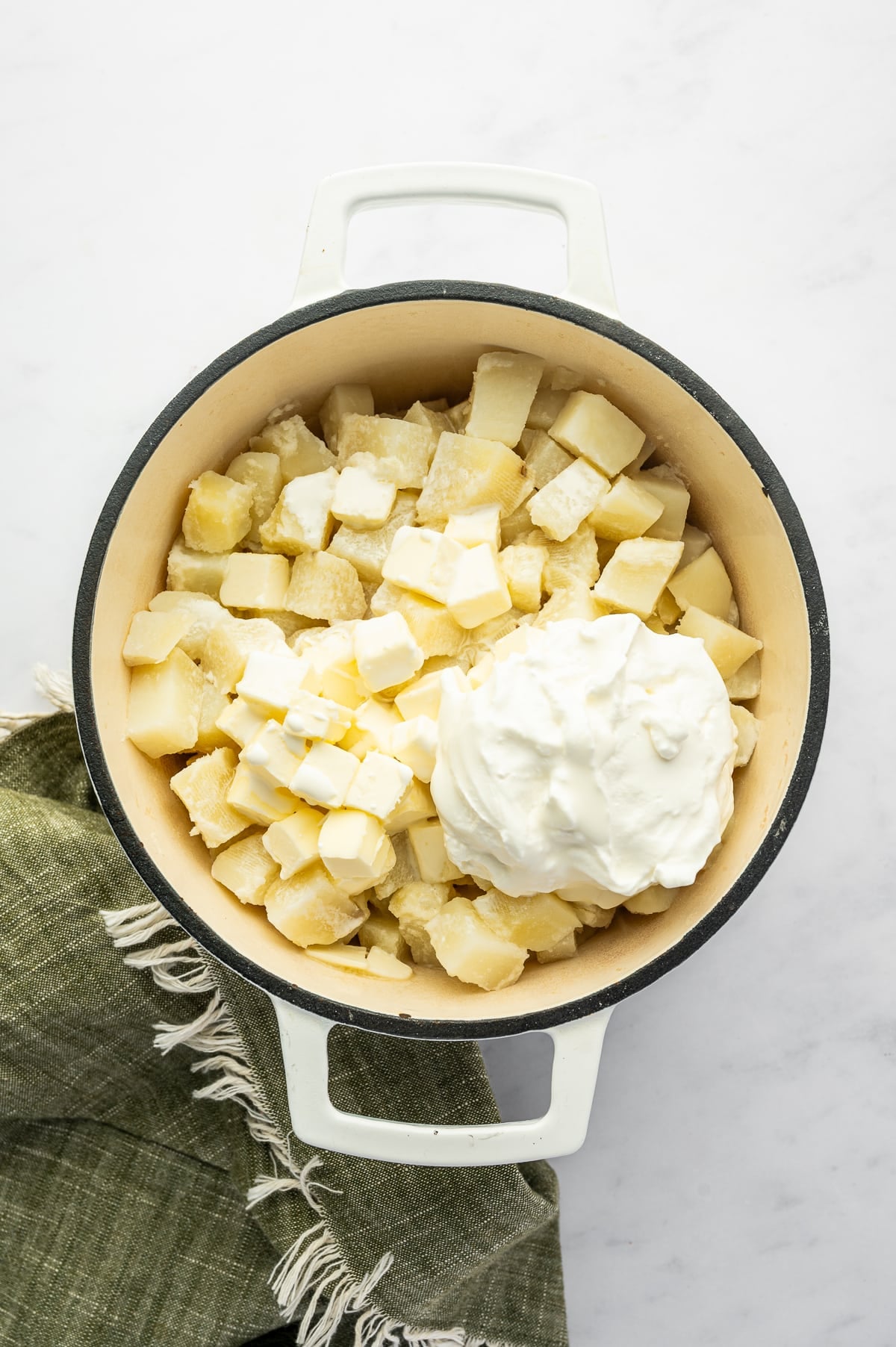 Overhead view of cooked Russet potatoes in a white enameled Dutch oven topped with cubes of pale yellow butter and a large spoonful of white sour cream. A dark green fringed towel sits beside the pot.