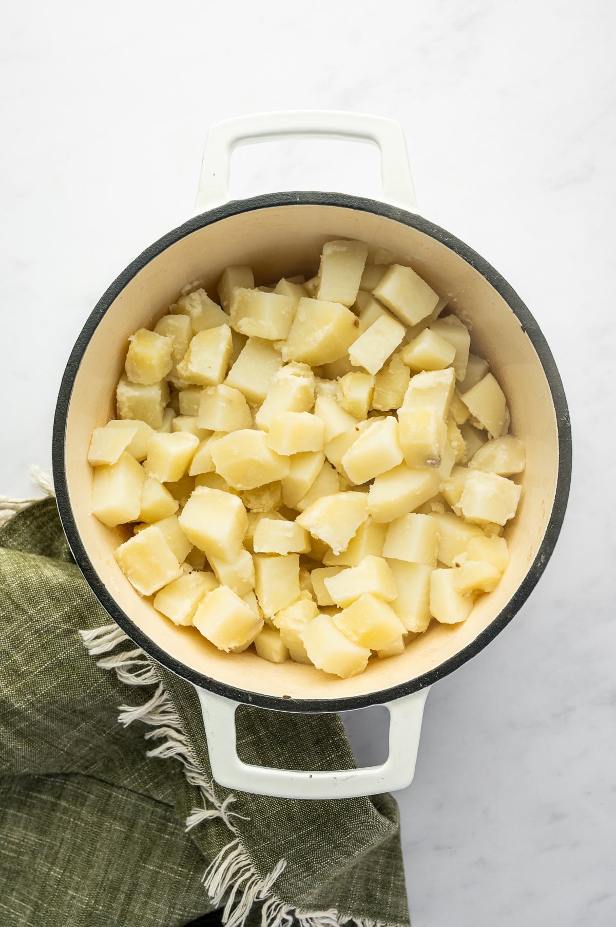 Overhead view of cooked Russet potato chunks drained in a white enameled Dutch oven with a cream interior. The potatoes look soft and dry, with a dark green fringed towel beside the pot.