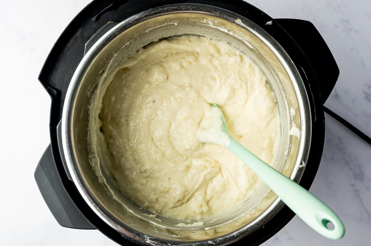 Thick, creamy mashed potatoes with soft swirls and a few small lumps fill the Instant Pot, with a pale green silicone spatula resting in the mixture.