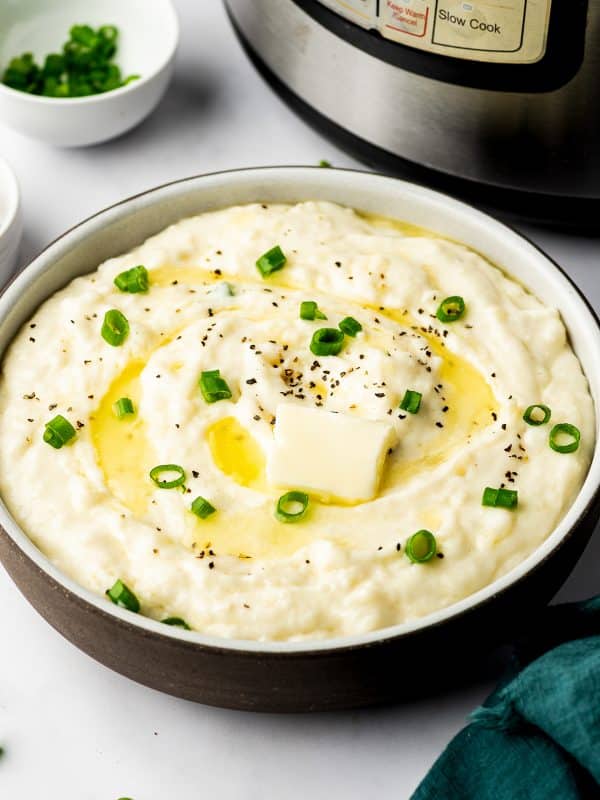 A wide ceramic bowl filled with creamy mashed potatoes topped with melting butter, sliced green onions, and black pepper, with an Instant Pot and dark teal napkin nearby.