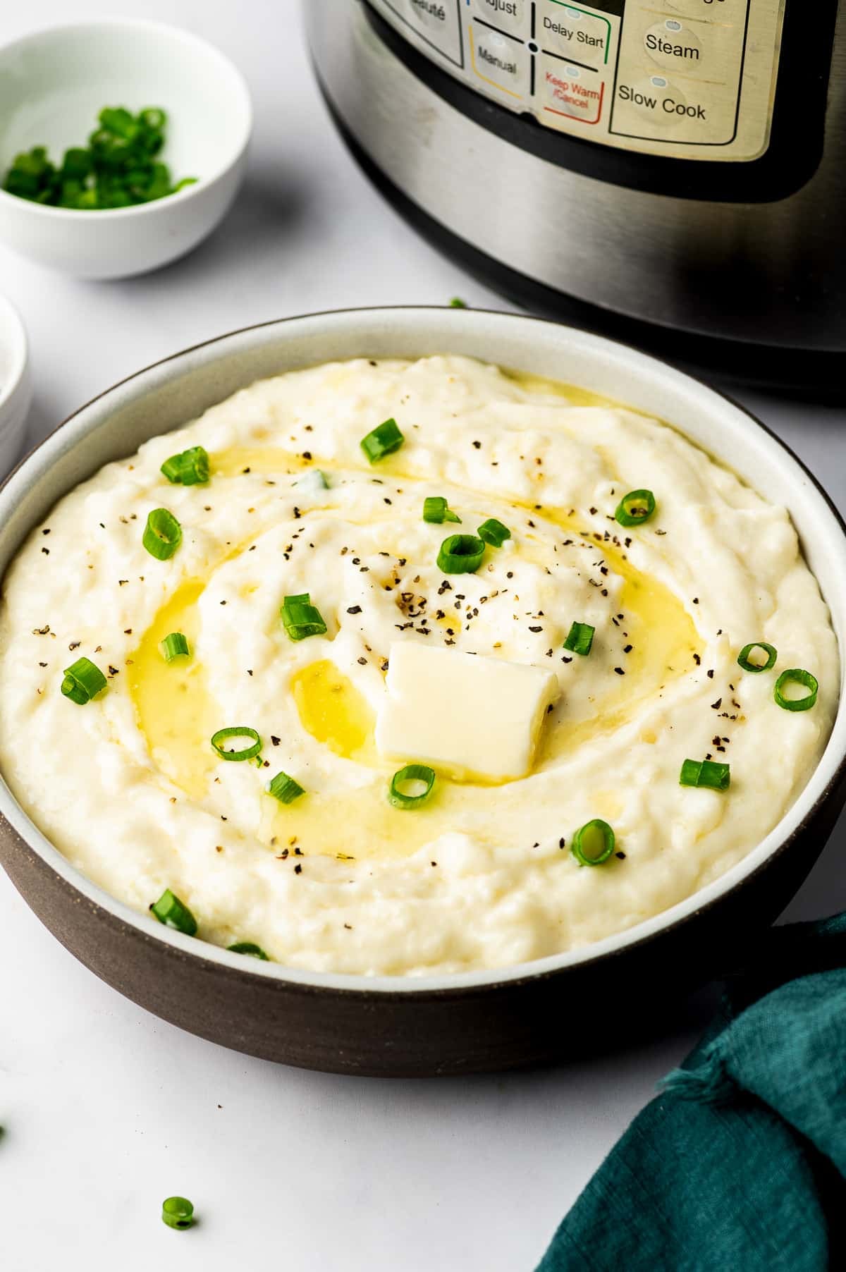 A wide ceramic bowl filled with creamy mashed potatoes topped with melting butter, sliced green onions, and black pepper, with an Instant Pot and dark teal napkin nearby.