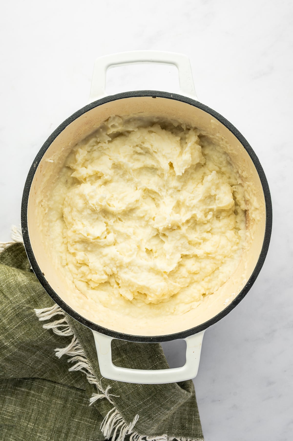 Overhead view of creamy mashed potatoes filling a white enameled Dutch oven with a cream-colored interior. The potatoes have a soft, fluffy texture with visible swirls from mashing.