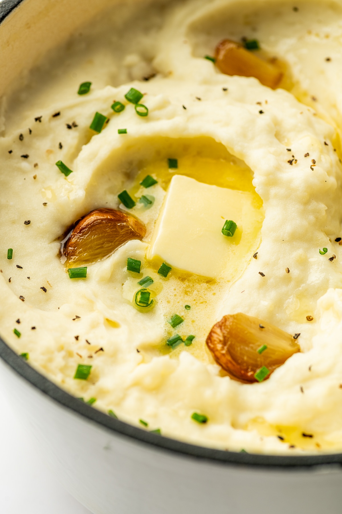 Close-up view of creamy roasted garlic mashed potatoes in a white enameled Dutch oven with a square pat of melting butter in the center, golden roasted garlic cloves, chopped green chives, and cracked black pepper.