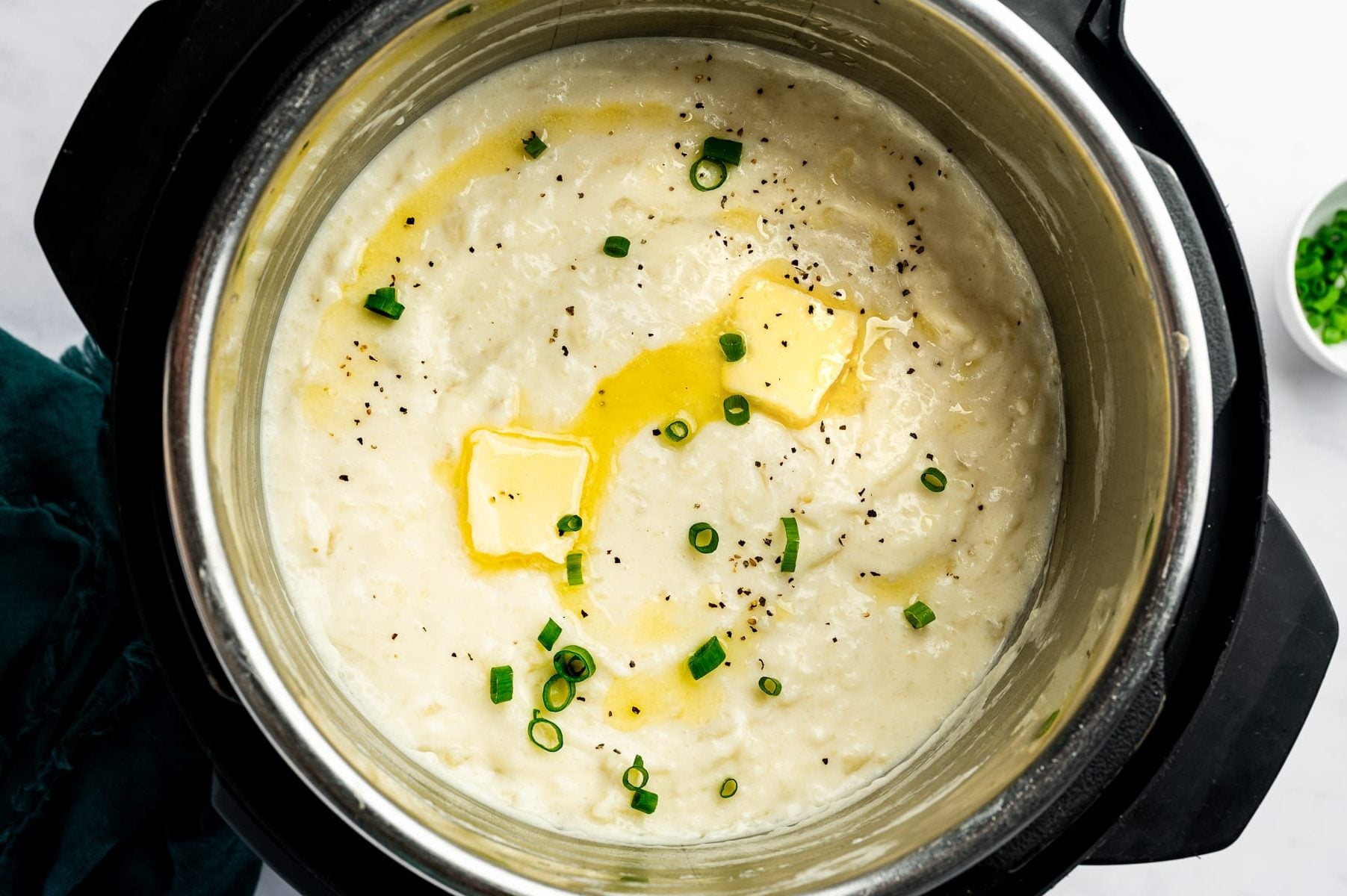 Creamy mashed potatoes inside a stainless steel Instant Pot, topped with two pieces of melting butter, chopped green onions, and freshly ground black pepper.