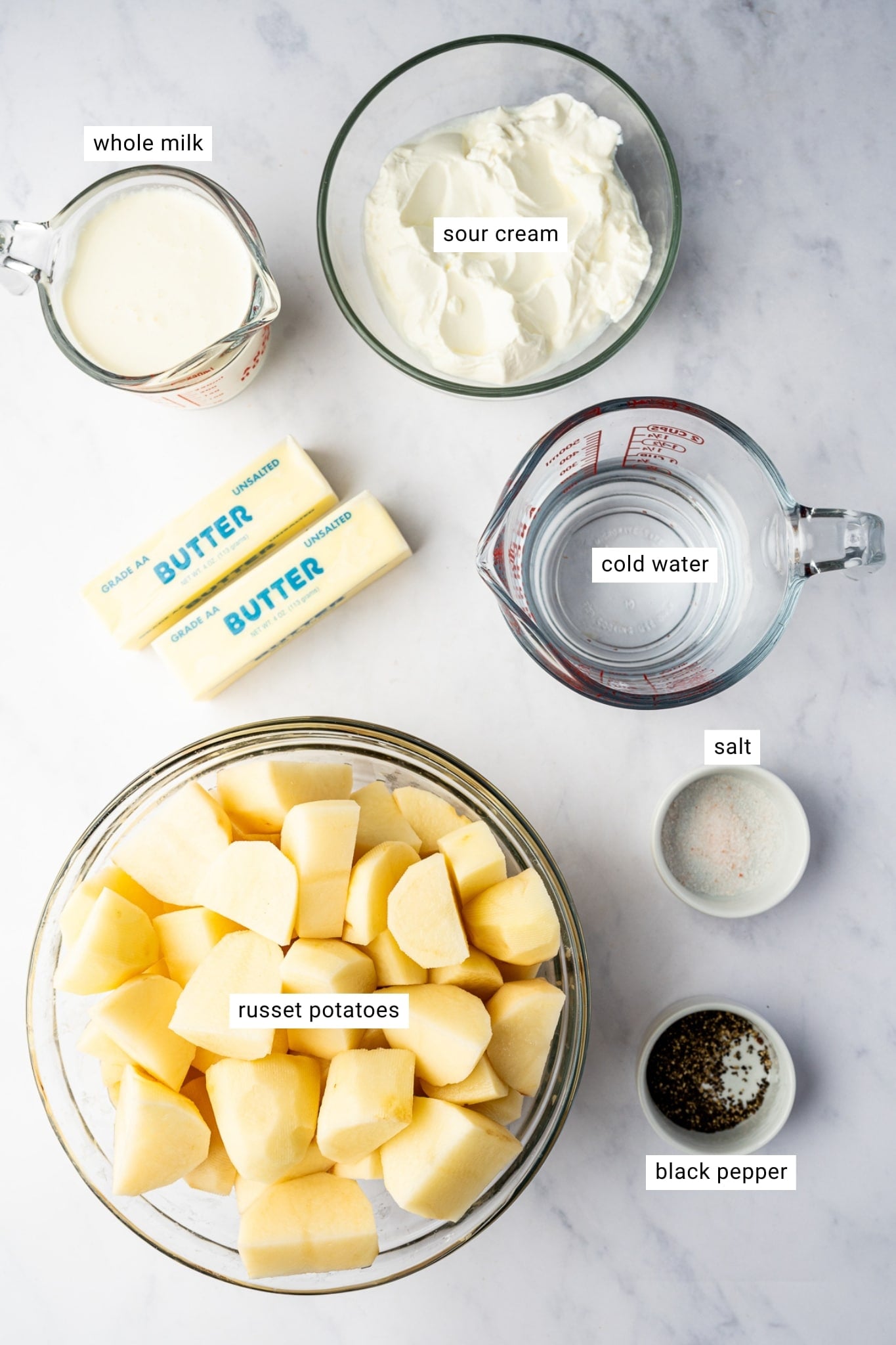 Labeled ingredients for Instant Pot mashed potatoes arranged on a white countertop, including peeled and chopped russet potatoes, two sticks of unsalted butter, half-and-half, sour cream, cold water, salt, and black pepper.