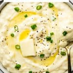Pinterest graphic featuring the words "Instant Pot Mashed Potatoes" above a bowl of thick, creamy mashed potatoes topped with melting butter, sliced green onions, and freshly ground black pepper. A gold spoon holds a generous scoop of mashed potatoes along the side of the bowl.