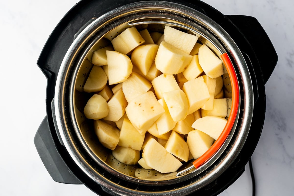 Peeled russet potatoes cut into large chunks and arranged in a metal steamer basket with an orange silicone handle inside an Instant Pot.