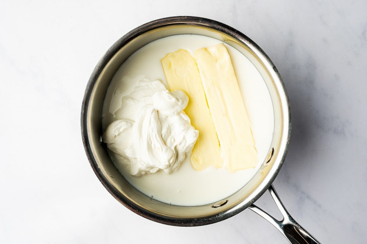 Two sticks of pale yellow butter and a generous scoop of thick white sour cream sit in a stainless steel saucepan filled with milk or cream.