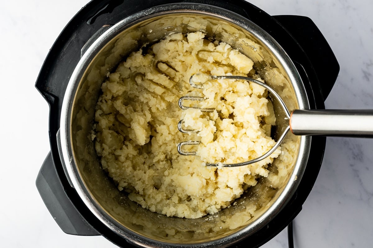 A stainless steel potato masher rests in a pot of partially mashed russet potatoes, with small, fluffy potato pieces and larger chunks still visible.