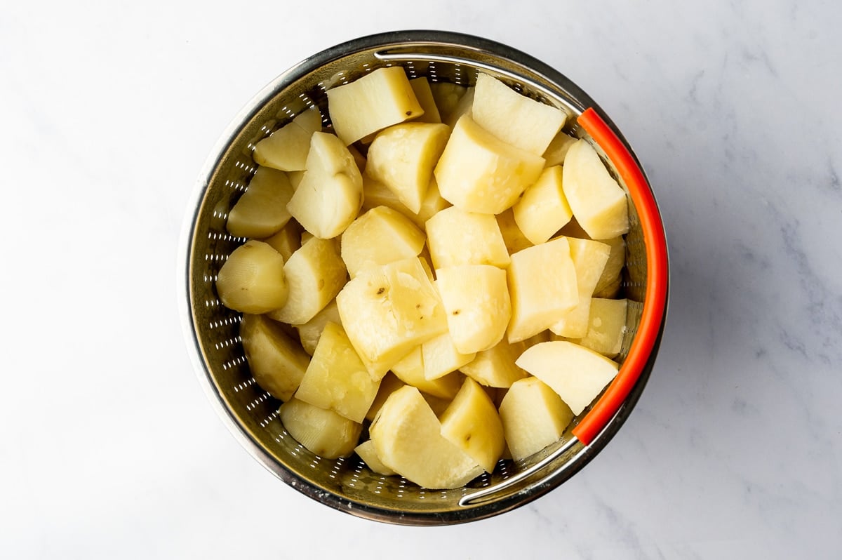 A round metal steamer basket filled with peeled russet potato chunks sits on a white countertop, with an orange silicone handle along one side.