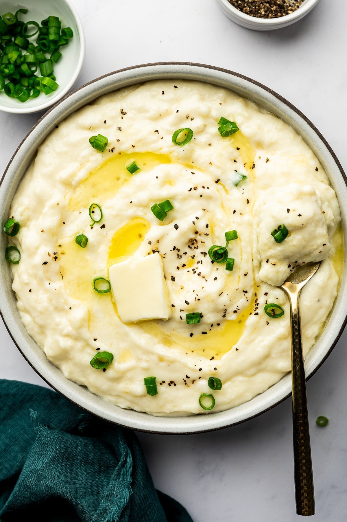 A large ceramic bowl filled with thick, creamy mashed potatoes, swirled with melted yellow butter and sprinkled with green onions and black pepper. A gold spoon holds a generous scoop of the fluffy potatoes.