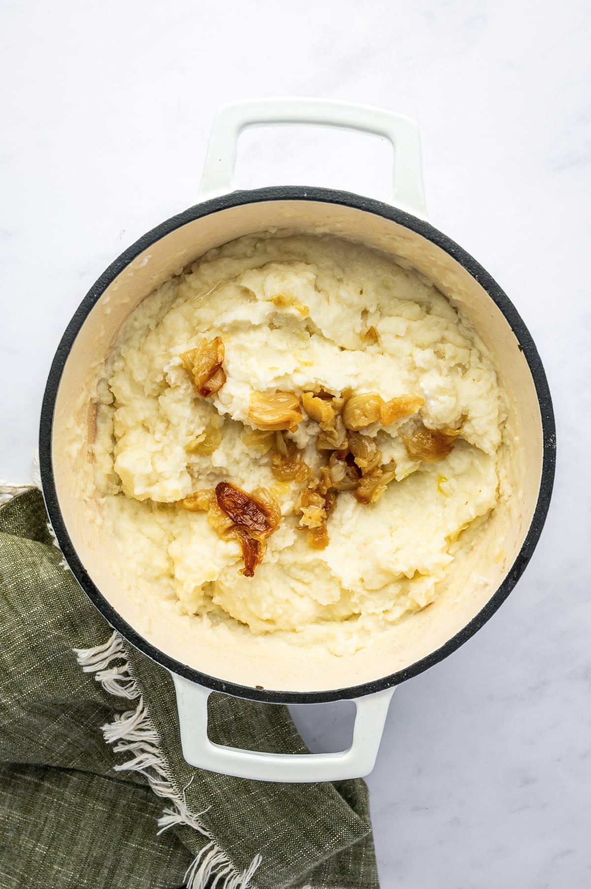 Overhead view of creamy mashed potatoes in a white enameled Dutch oven topped with pieces of golden roasted garlic. A dark green fringed kitchen towel is visible beside the pot.