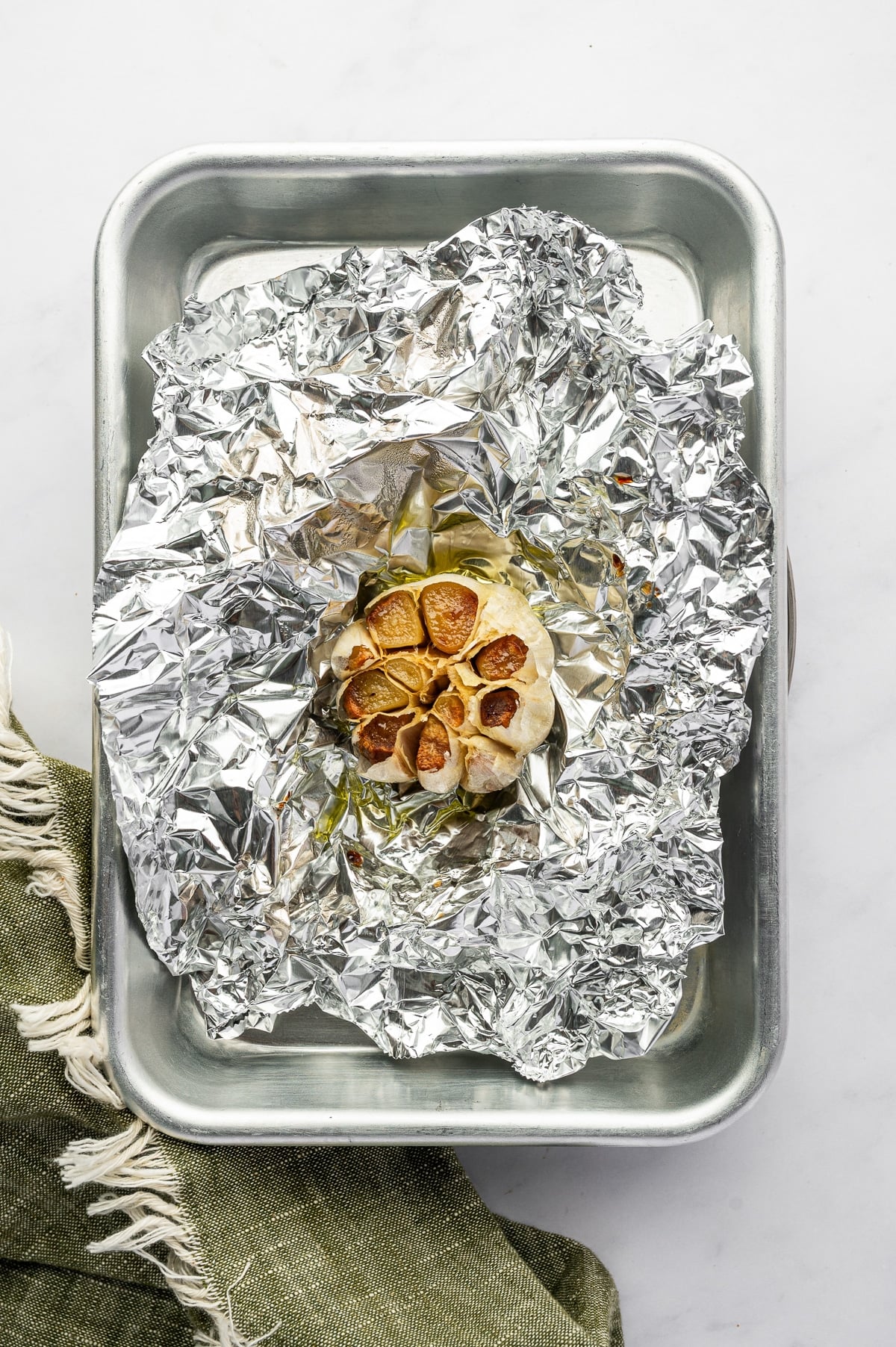 Overhead view of a roasted garlic bulb opened on crinkled aluminum foil inside a silver metal baking pan. The exposed garlic cloves are deeply golden brown and caramelized, with a dark green fringed towel along the side.