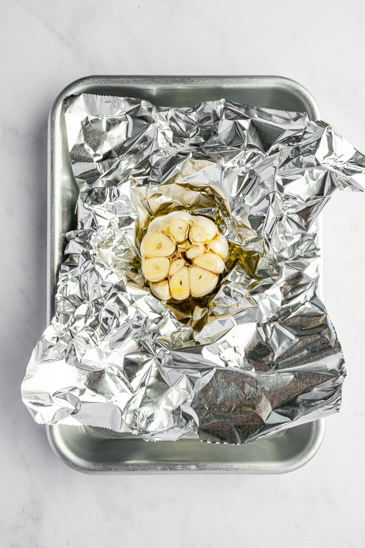 Overhead view of a whole garlic bulb with the top sliced off to expose the cloves, drizzled with olive oil and resting on crinkled aluminum foil inside a silver metal baking pan on a white surface.