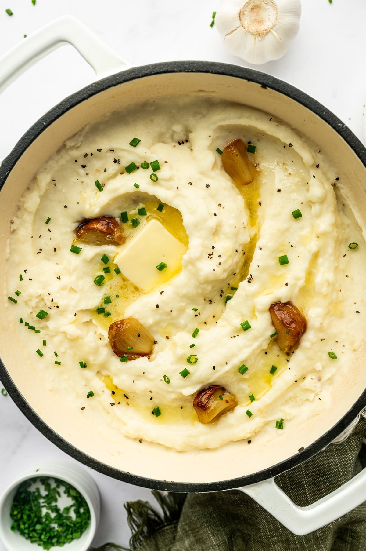 Overhead view of creamy roasted garlic mashed potatoes in a white enameled Dutch oven, topped with a pat of melting butter, whole roasted garlic cloves, chopped fresh chives, and cracked black pepper. A whole garlic bulb and small bowl of chives sit nearby.