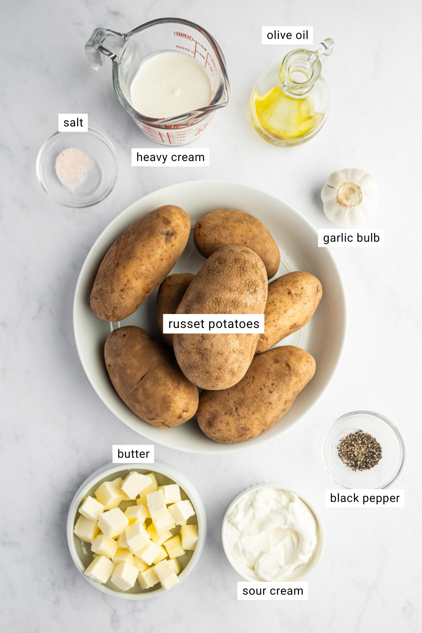 Overhead ingredient layout for roasted garlic mashed potatoes showing Russet potatoes in a white bowl, cubed butter, sour cream, heavy cream, olive oil, a whole garlic bulb, salt, and black pepper on a white marble surface. Text labels identify "russet potatoes," "butter," "sour cream," "heavy cream," "olive oil," "garlic bulb," "salt," and "black pepper."