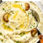 Vertical Pinterest image showing a close-up of creamy roasted garlic mashed potatoes in a white serving bowl topped with a pat of melted butter, browned roasted garlic cloves, chopped green chives, and black pepper. A white text box at the top reads "Roasted Garlic Mashed Potatoes" in black lettering with a yellow line between the two lines of text. "THEFORKEDSPOON.COM" appears along the bottom.