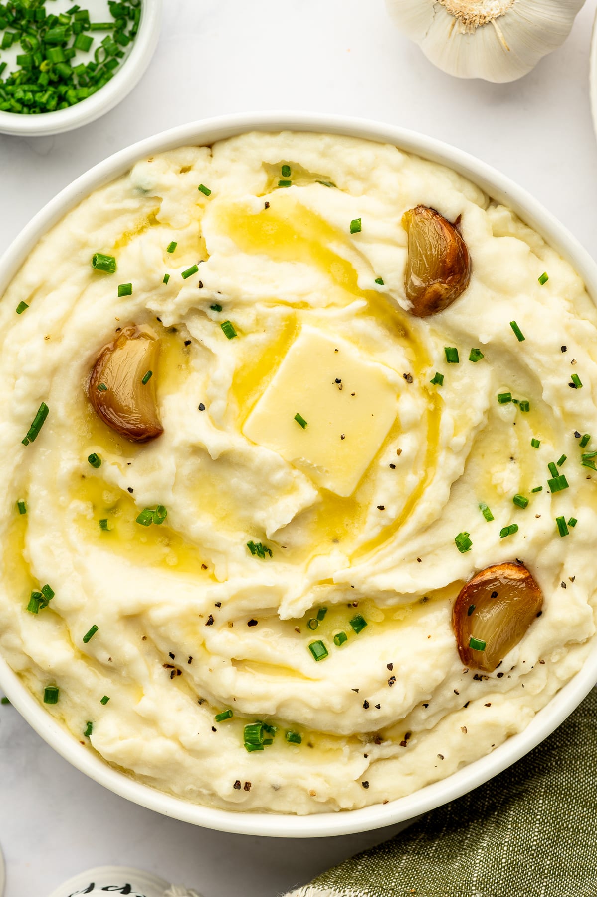 Overhead close-up of roasted garlic mashed potatoes served in a round white bowl, topped with a square pat of melting butter, golden roasted garlic cloves, chopped green chives, and cracked black pepper. A whole garlic bulb and bowl of chives sit around the serving dish.