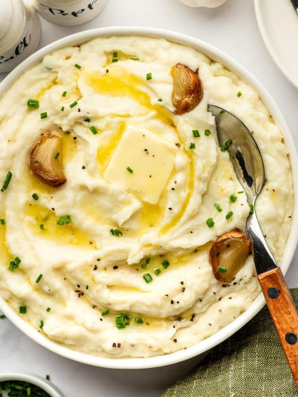 Overhead view of creamy roasted garlic mashed potatoes in a round white serving bowl with melted butter, roasted garlic cloves, chopped chives, and black pepper. A metal serving spoon with a wooden handle rests inside the bowl, with white salt and pepper shakers and a garlic bulb nearby.