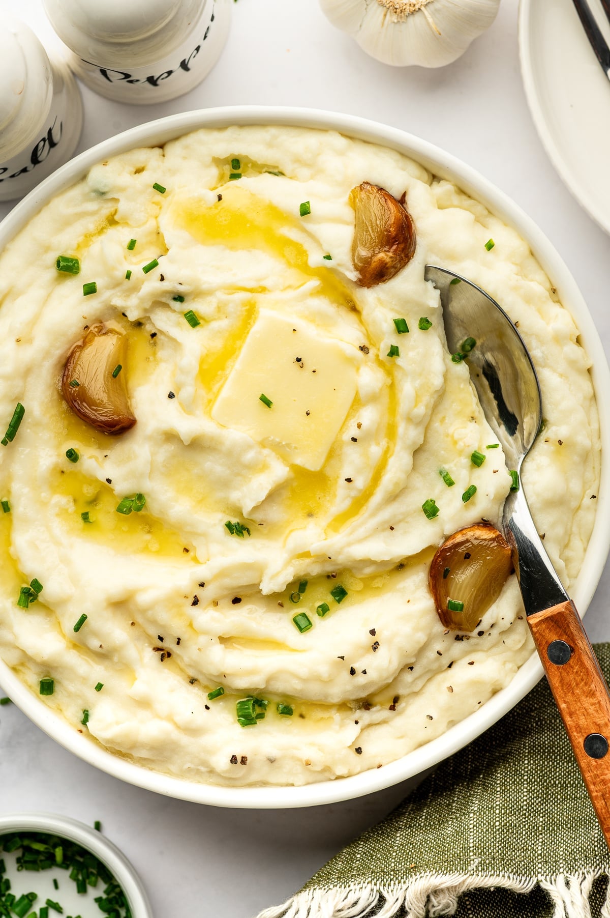 Overhead view of creamy roasted garlic mashed potatoes in a round white serving bowl with melted butter, roasted garlic cloves, chopped chives, and black pepper. A metal serving spoon with a wooden handle rests inside the bowl, with white salt and pepper shakers and a garlic bulb nearby.