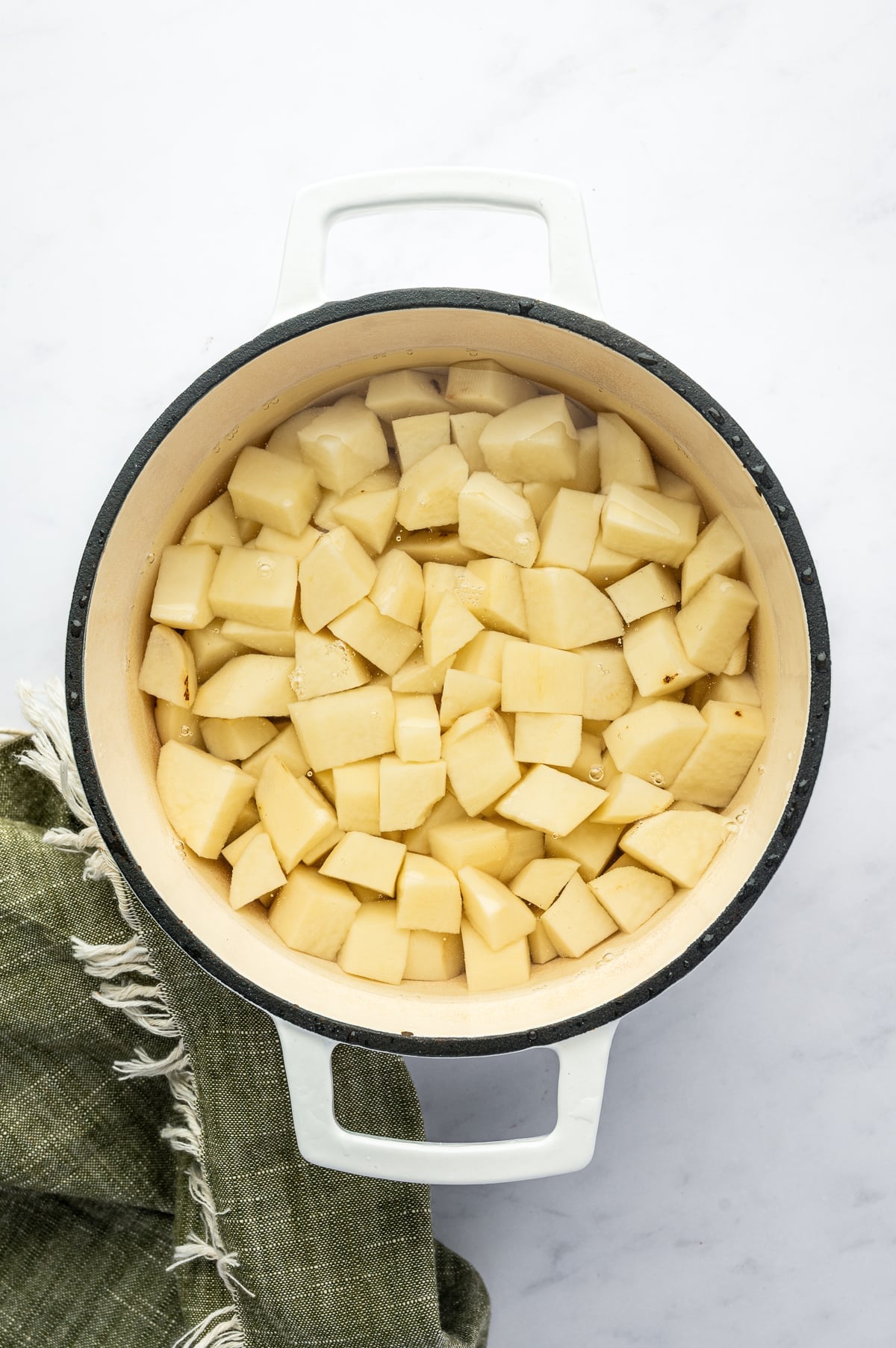 Overhead view of peeled Russet potatoes cut into chunks and submerged in cold water inside a white enameled Dutch oven with a cream-colored interior. A dark green fringed kitchen towel rests beside the pot.