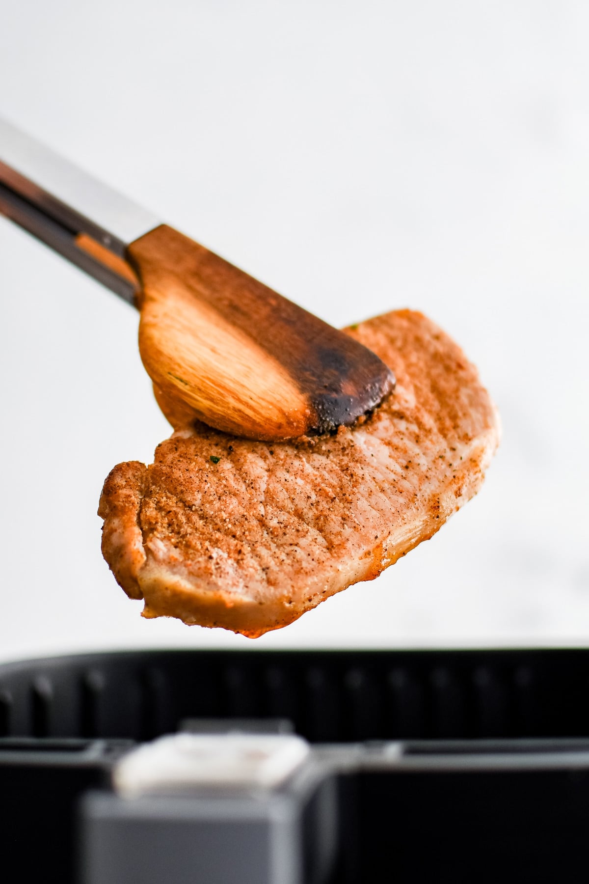 Tongs removing one fully cooked boneless pork chop from the basket of an air fryer.