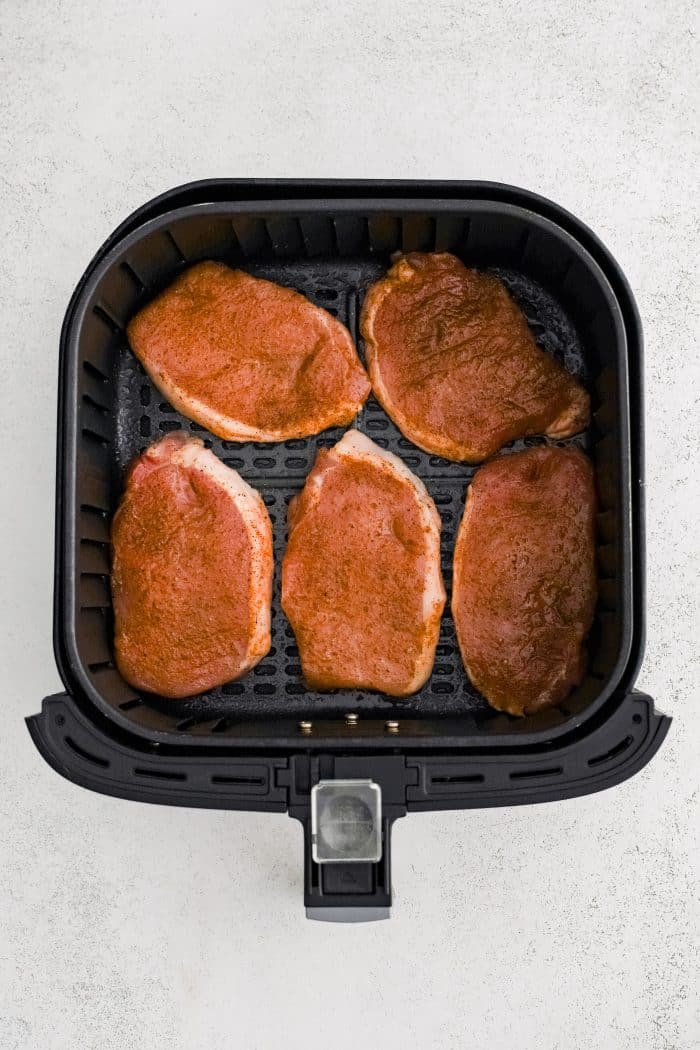 Five boneless raw pork chops seasoned with paprika, salt, pepper, onion powder, and garlic powder, in the basket of an air fryer.