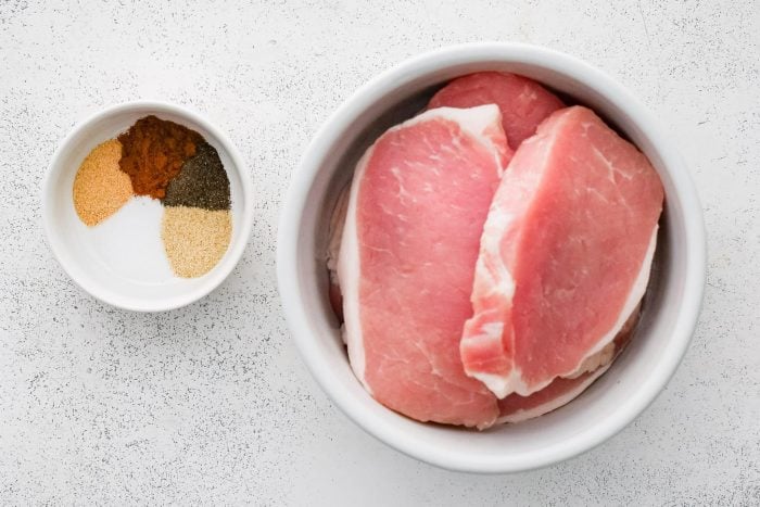 One larger white bowl filled with boneless pork chops and one small white bowl filled with various seasonings.