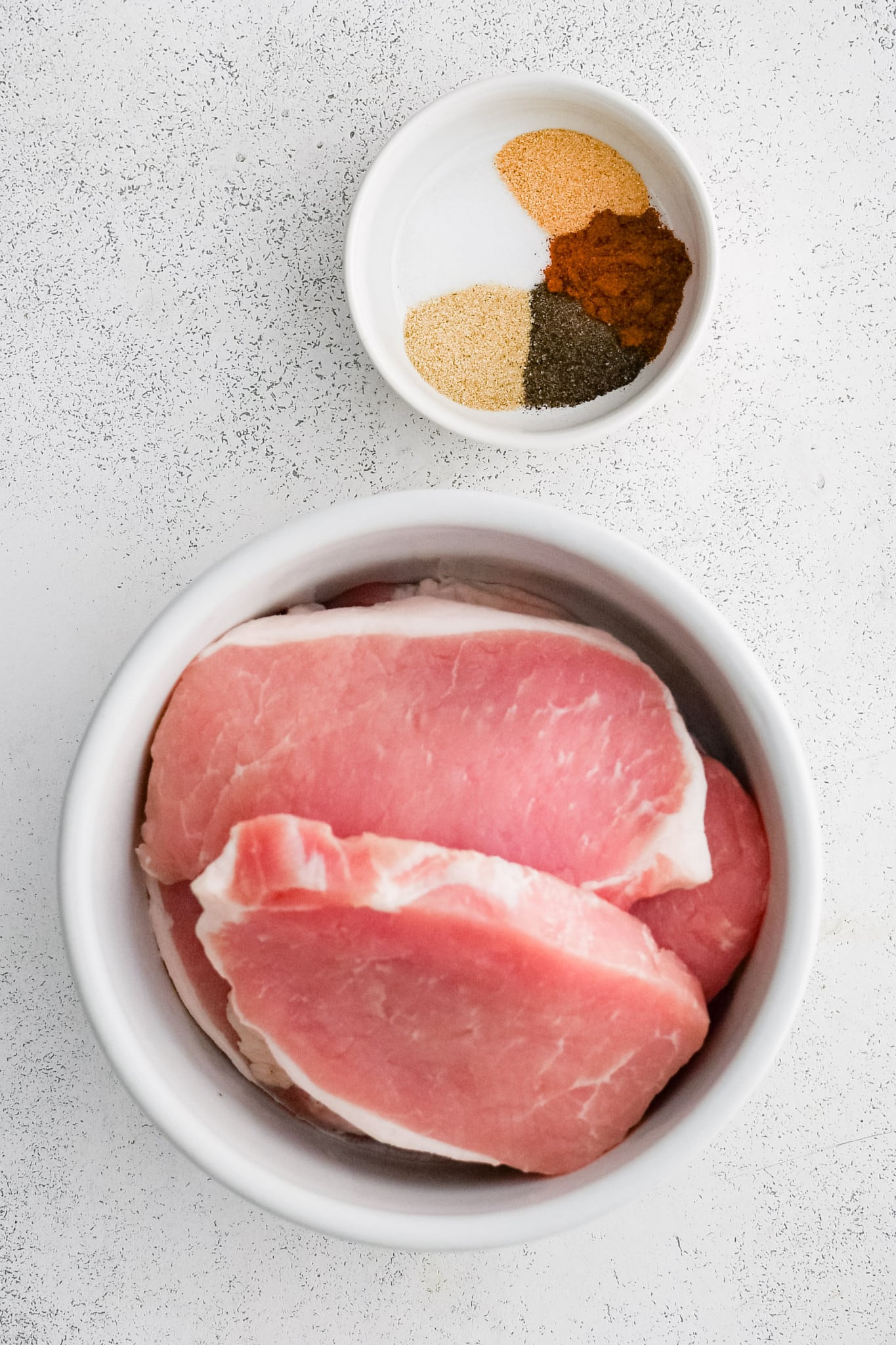 One larger white bowl filled with boneless pork chops and one small white bowl filled with various seasonings.