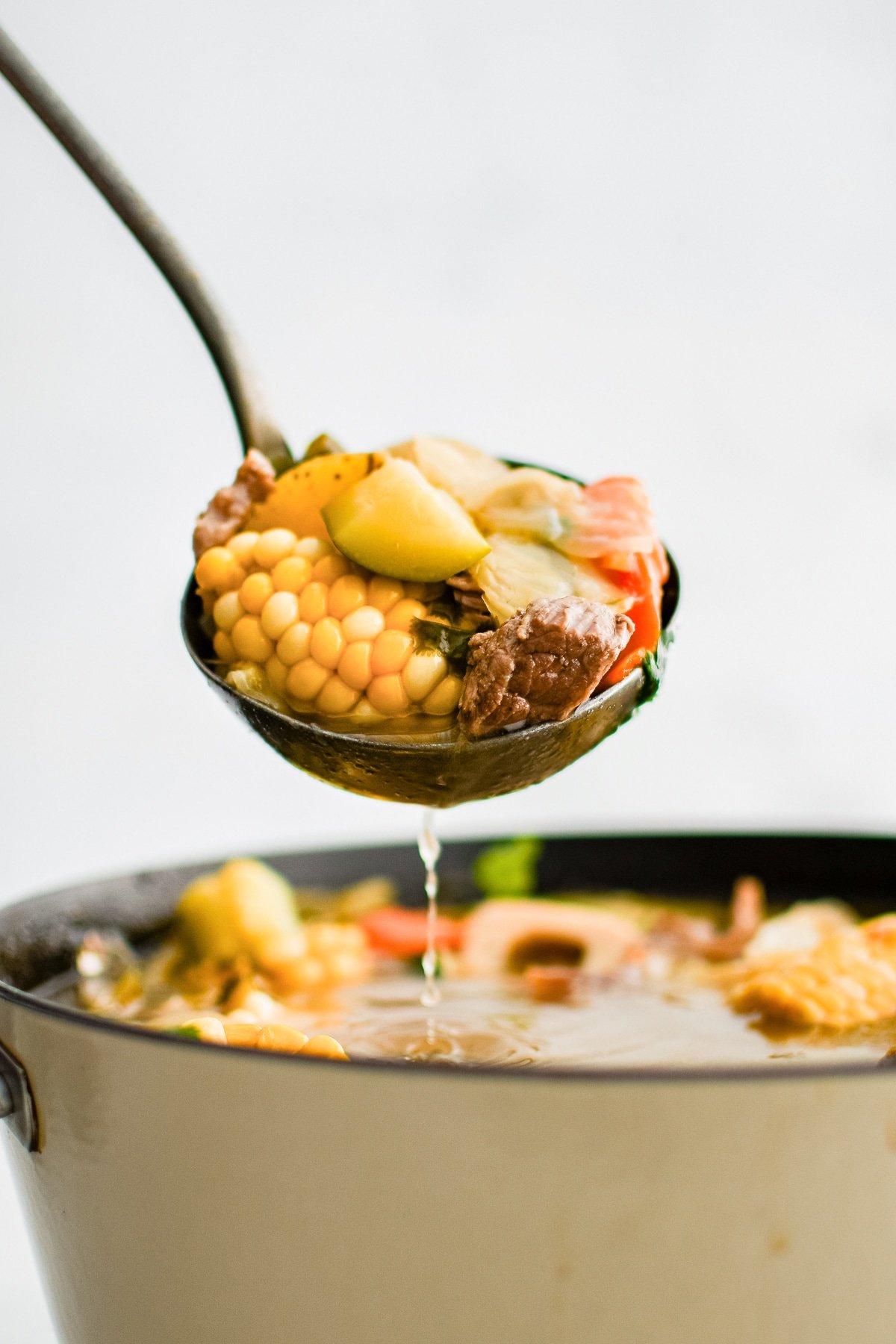 Ladle filled with caldo de res with chunks of corn, zucchini, potato, and carrot.