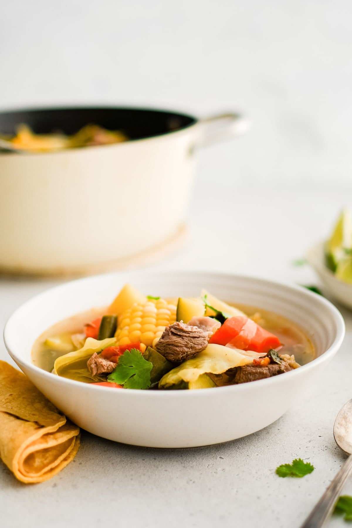 White soup bowl filled with Mexican beef and vegetable soup with tender chunks of beef and tender cooked vegetables like zucchini, potatoes, carrots, and corn.