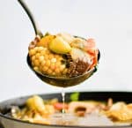 Ladle filled with caldo de res with chunks of corn, zucchini, potato, and carrot.