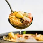 Ladle filled with caldo de res with chunks of corn, zucchini, potato, and carrot.