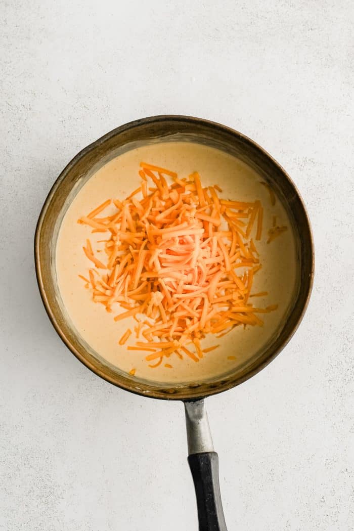 Shredded cheddar cheese added to a medium pot filled with thickened milk sauce.