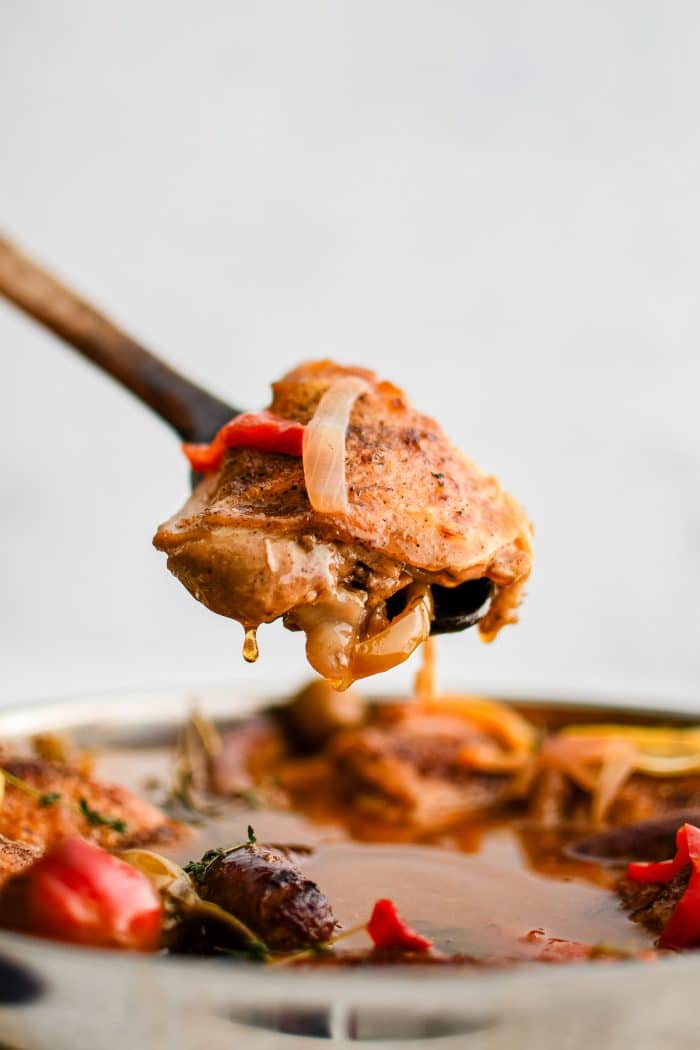 Wooden spatula filled with a braised bone-in skin-on chicken thigh with peppers and onions in sauce.