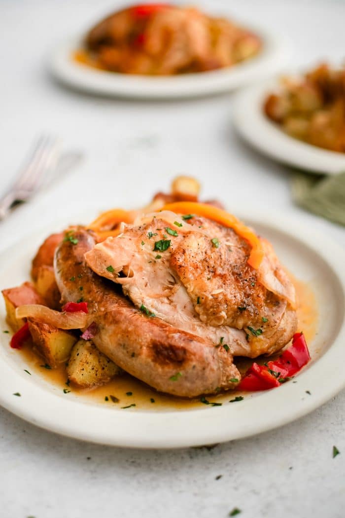 White plate filled with a crispy braised chicken thigh and whole cooked Italian sausage smothered in broth and served with roasted potatoes.