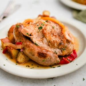 White plate filled with a crispy braised chicken thigh and whole cooked Italian sausage smothered in broth and served with roasted potatoes.