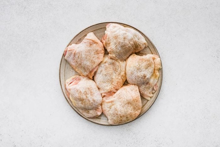 Six raw bone-in skin-on chicken thighs seasoned with salt and pepper on a plate.